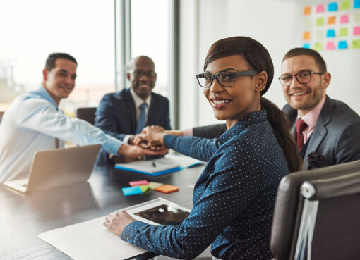 Successful African American team leader turning to smile at the camera as her multiracial team of executives links hands across the table