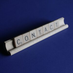 Wooden Scrabble tiles spelling the word CONTACT on a letter rack against a blue background