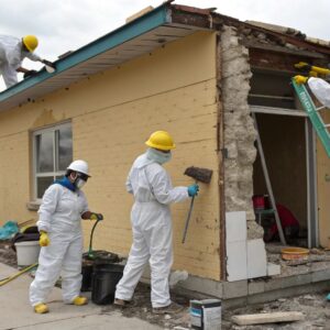 Restoration crew wearing protective suits and helmets repairing the exterior of a damaged building, removing debris and working on structural restoration.